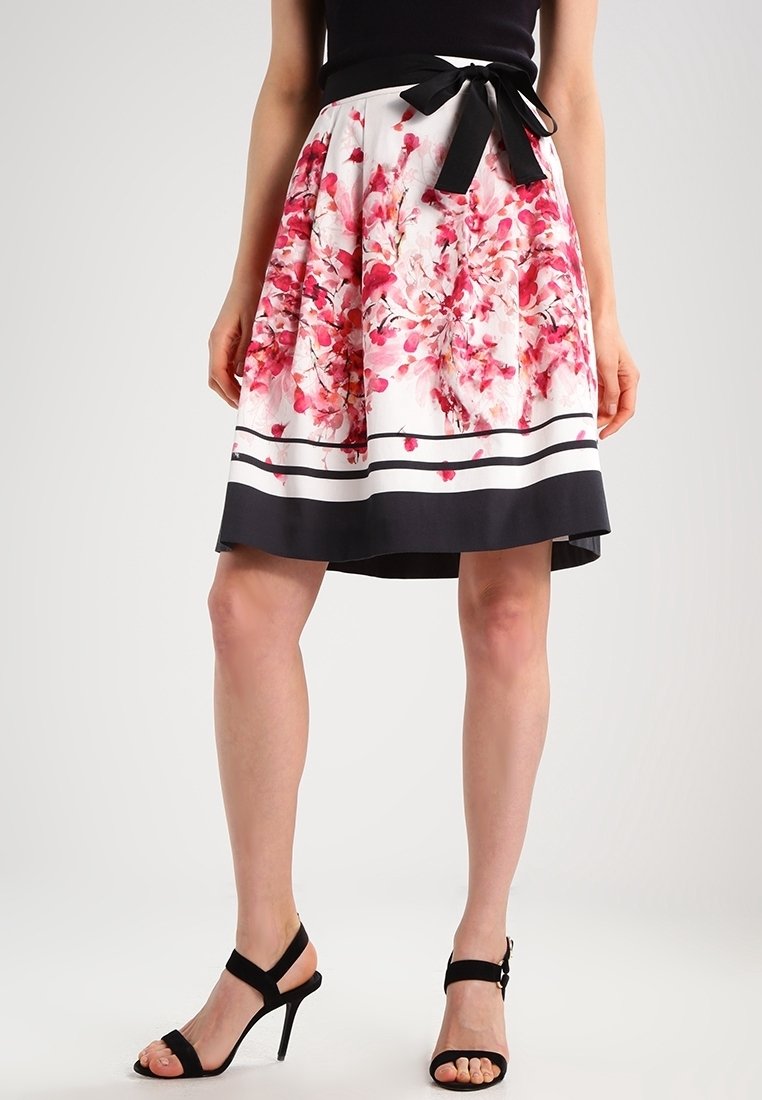 Jupe à imprimé floral avec une base blanche, présentant des motifs de fleurs roses, une taille noire et un ourlet foncé avec des rayures. Accent en noeud noir.