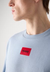 Svetlo modrý sweatshirt s okrúhlym výstrihom, ktorý má výrazný červený obdĺžnikový https://twilightmuse.com/v0/patch s čiernym vyšitým textom "HUGO".