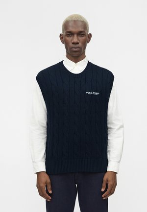 Mand med kort blondt hår iført en navy blå kabelstrik ærmeløs sweater over en hvid skjorte med knapper, stående mod en ensfarvet hvid baggrund.
