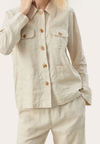 Person iført en beige linned skjorte med knapper og påsyede lommer samt matchende bukser, poserer med den ene hånd på halsen og den anden i lommen.