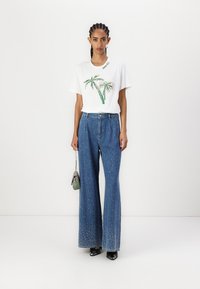 Witte t-shirt met een groen en zilver palmboomontwerp, gecombineerd met hoogvergde blauwe denim wijdlopende pantalons met sprankelende accenten. Kleine handtas.