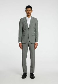 Completo grigio in tessuto testurizzato, vestibilità aderente, giacca monopetto con rever a punte, abbinato a una camicia bianca e scarpe nere.