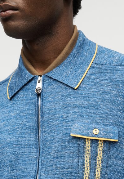 Κοντινό πλάνο ατόμου που φοράει μπλε πλεκτό πουκάμισο με φερμουάρ και κίτρινες λεπτομέρειες, τσέπη με κουμπιά στο στήθος και εμφανές γιακά σε μπεζ εσωτερικό πουκάμισο.