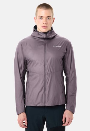 Giovane uomo con indosso una giacca con cappuccio Vaude viola chiaro con cerniera frontale e pantaloni neri, in piedi davanti a uno sfondo neutro.