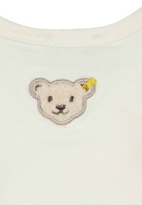 T-shirt en tissu blanc arborant un patch en relief brodé représentant un visage d'ours, avec un accent jaune sur l'oreille droite et des détails en contour gris.