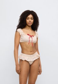 Σετ μαγιό δύο τεμαχίων σε ανοιχτό ροδακινί με κόκκινες πουά. Η μπλούζα έχει τετράγωνη λαιμόκοψη και φουσκωτά μανίκια· το κάτω μέρος διαθέτει βολάν στο τελείωμα.