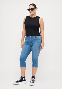 Top crop negro sin mangas combinado con jeans capri de denim azul claro. La modelo lleva zapatillas altas negras y gafas de sol. Diseño sencillo, forma ajustada.