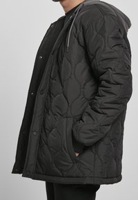Personne portant une veste matelassée noire avec capuche, poche latérale, boutons-pression et main partiellement dans la poche.