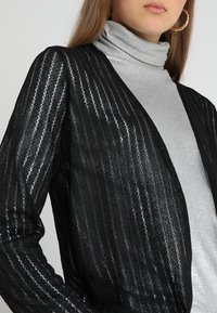 Černý kardigan z průhledné, texturované látky s vertikálními pruhy, nošený přes stříbrný rolák. Výrazný doplněk: náušnice kruhy.