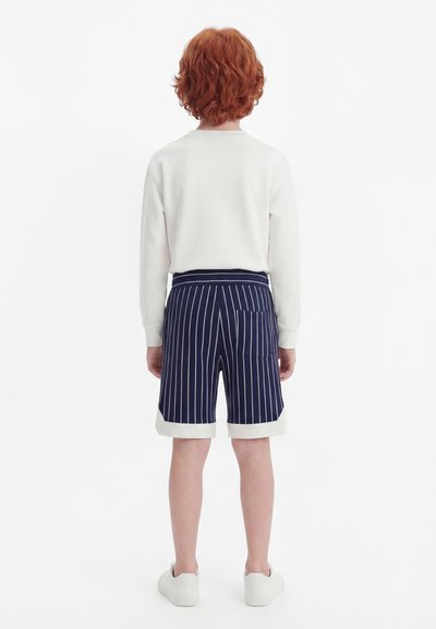 Niño con cabello rizado y rojo de espaldas, vestido con una camiseta blanca de manga larga, pantalones cortos azul marino a rayas con puños blancos y zapatillas blancas.