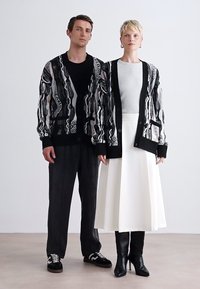 Two models stand side by side. Each wears a patterned black-and-white cardigan; one has a grey top, white skirt, and black boots, the other a black shirt and pants.