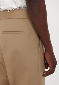 Gros plan sur la main d'une personne à côté d'un pantalon beige sur mesure avec une poche passepoilée et un haut en dentelle blanche.