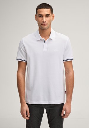 Mężczyzna w białej koszulce polo z krótkim rękawem, z granatowymi paskami na mankietach rękawów i czarnych dżinsach, stoi na prostym tle.