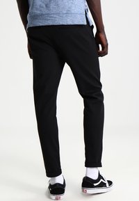 Pantalon noir fuselé avec une texture lisse, doté de poches latérales et d'une taille mi-haute, accompagné de baskets noires et de chaussettes blanches.