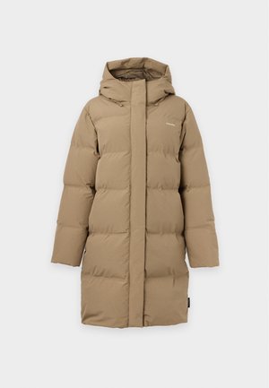 Femme portant une doudoune à capuche beige, un pull gris, un pantalon noir à jambes larges et des chaussures noires, debout sur un fond blanc uni.