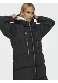 Gilet rembourré noir avec capuche, présentant un design matelassé, des fermetures éclair argentées et une doublure douce en fausse fourrure. Manches en tricot vert texturé.