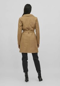 Abrigo color camel con cinturón, cuello de pico y diseño ajustado. Combinado con pantalones negros de corte slim y botas altas hasta la rodilla. Textura de tela suave.