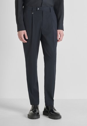Man in op maat gemaakte donkerblauwe broek en zwarte leren veterschoenen, staand tegen een effen lichte achtergrond.
