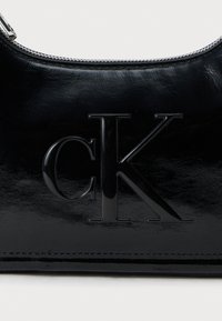 Čierna lakovaná kožená kabelka so zipsovým uzáverom a embosovaným logom "CK" v strede. Hladký povrch a lesklý vzhľad.