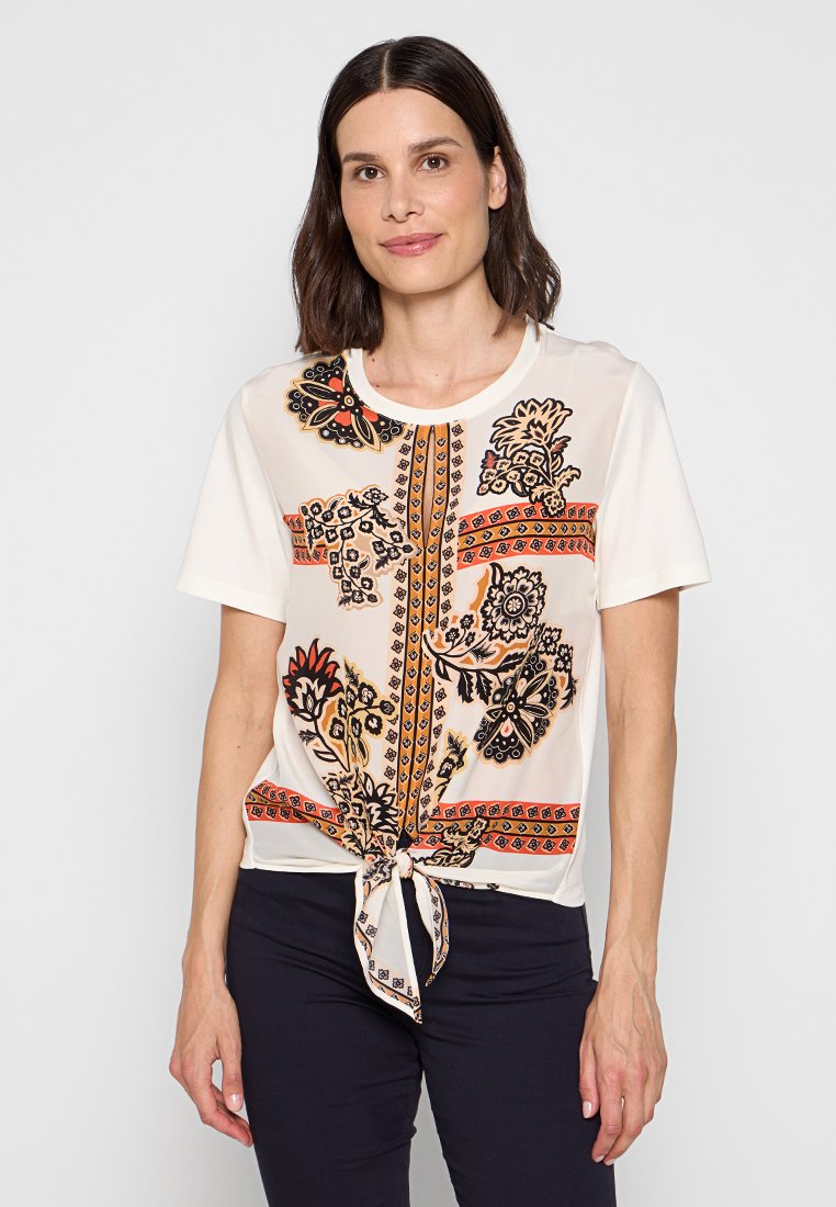Korte mouwen blouse in crème met zwarte en oranje bloemenpatronen. Heeft een ronde hals en een knoopdetail aan de onderkant. Zachte stoftextuur.