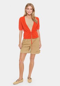 Rode gebreide cardigan met korte mouwen, met een strik in de taille, gecombineerd met beige shorts. Model draagt geweven instapschoenen.