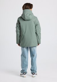 Green waterproof jacket with a hood, long sleeves, and a relaxed fit. Paired with light blue jeans and black and white sneakers.