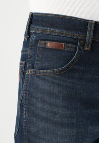 Vaqueros de mezclilla azul oscuro con una textura suave, costuras visibles y un parche de cuero de la marca en el bolsillo trasero. Diseño de cintura media.