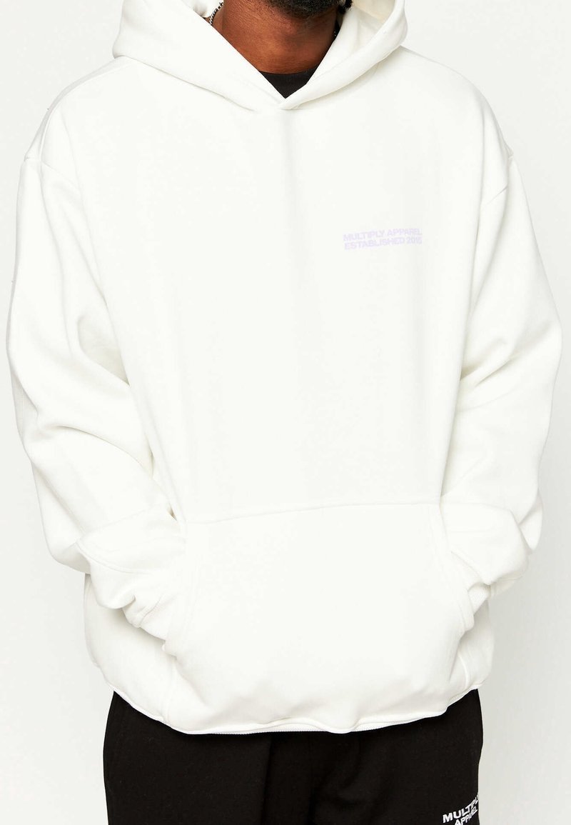 Homme portant un sweat à capuche blanc oversize avec les mains dans la poche avant et un pantalon noir sur fond uni.