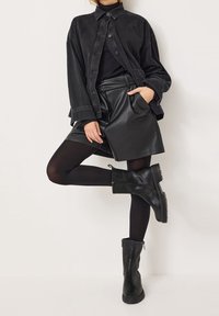 Chaqueta de mezclilla negra superpuesta sobre una blusa negra de canalé, combinada con una falda corta de cuero negro de cintura alta y botines. Manga sin mangas, detalle de bolsillo.