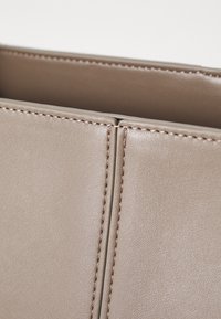 Beige nahkakäsilaukku, jossa on sileä pinta ja minimalistinen muotoilu, samoin kuin näkyvät tikit saumoissa ja suorat reunat.