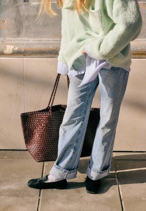 Person trägt einen hellgrünen Pullover, blaue aufgekrempelte Jeans, schwarze Schuhe, hält eine geflochtene braune Tragetasche und steht auf dem Bürgersteig neben einer Steinmauer.