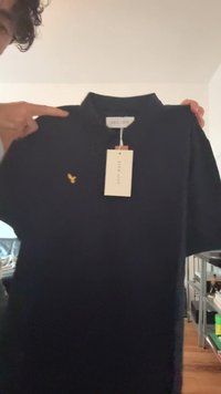 Eine Person hält ein schwarzes Polohemd mit einem gelben Vogel-Logo und einem weißen Etikett mit der Aufschrift "BLUE ONE" in der Nähe eines Raumes mit Regalen und einem Sofa.