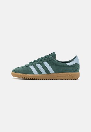 Zielony zamszowy sneaker z jasnoniebieskimi paskami i pętelką na pięcie, beżowa gumowa podeszwa oraz sznurowane zapięcie, widoczny od wewnętrznej strony.