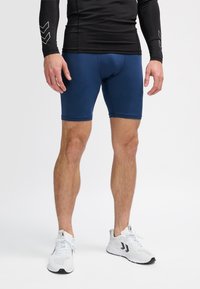 Man klädd i marinblå sportshorts, svart långärmad tröja och vita löparskor, ståendes på vit bakgrund.