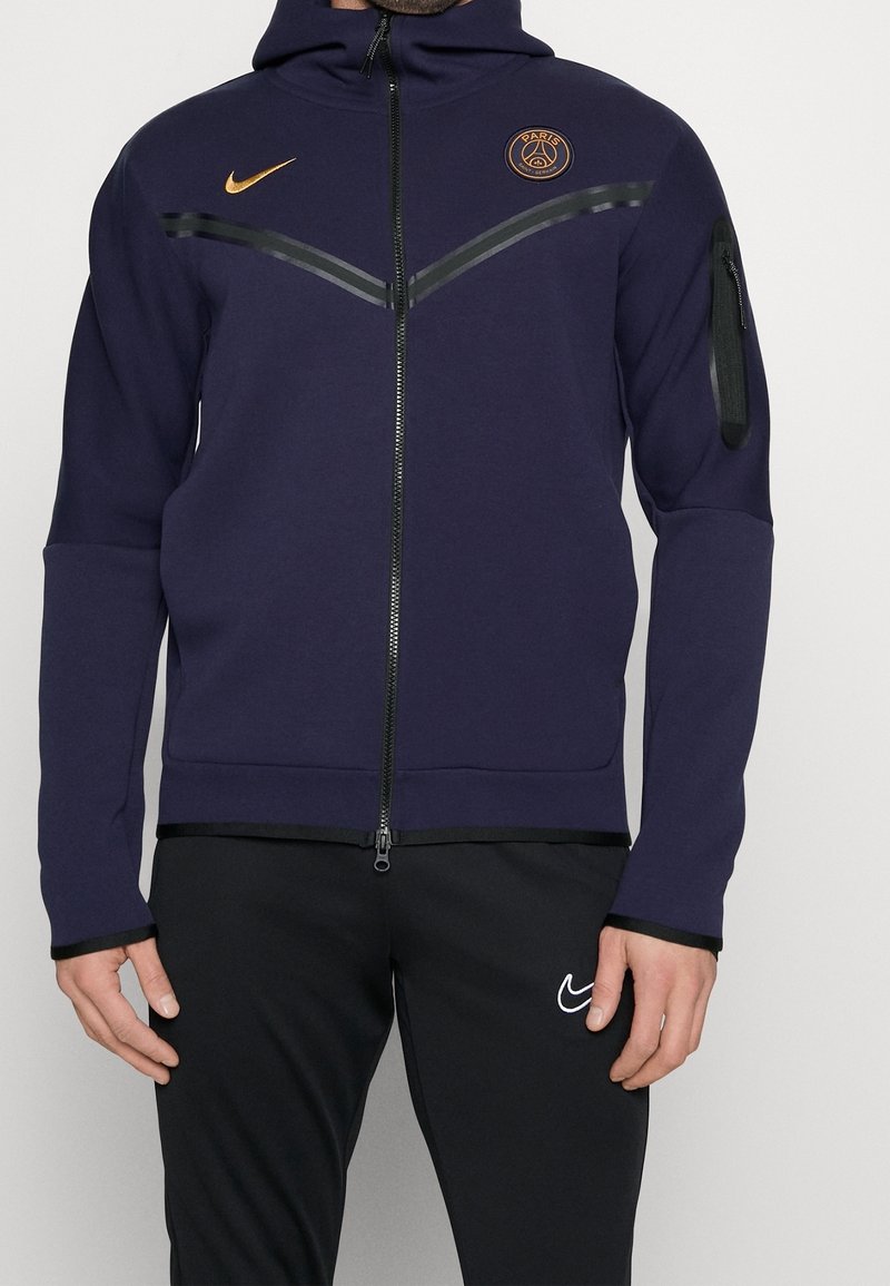 Homme portant un sweat à capuche zippé bleu marine Paris Saint-Germain et un pantalon Nike noir avec un logo Swoosh blanc sur la cuisse gauche.