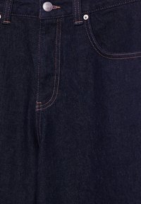 Mørkeblå denimjeans tæt på, der viser frontknap, lynlås, bæltestropper og frontlomme med orange stingdetaljer.