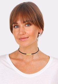 Sort choker halskæde med en glat guldfarvet metalaccent, båret om halsen. Simpelt design, flad tekstur, minimalistisch stil.