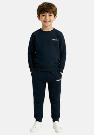 Jeune garçon souriant, portant un sweat-shirt bleu marine ellesse et un pantalon de jogging assorti avec des baskets blanches, les mains dans les poches.