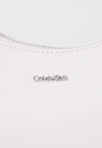 Calvin Kleinin vaaleanpunainen nahkalaukku hopeisilla metalliosilla, jossa on teksturoitu pinta ja vetoketjusuljenta yläreunassa.