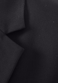 Gros plan sur un tissu noir montrant la texture et les bords pliés d’un vêtement, probablement un blazer ou une veste.
