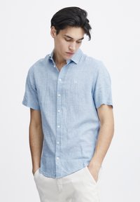 Camicia a maniche corte di colore blu chiaro con colletto a bottoni, realizzata in tessuto traspirante, con un piccolo logo sul lato sinistro del petto.