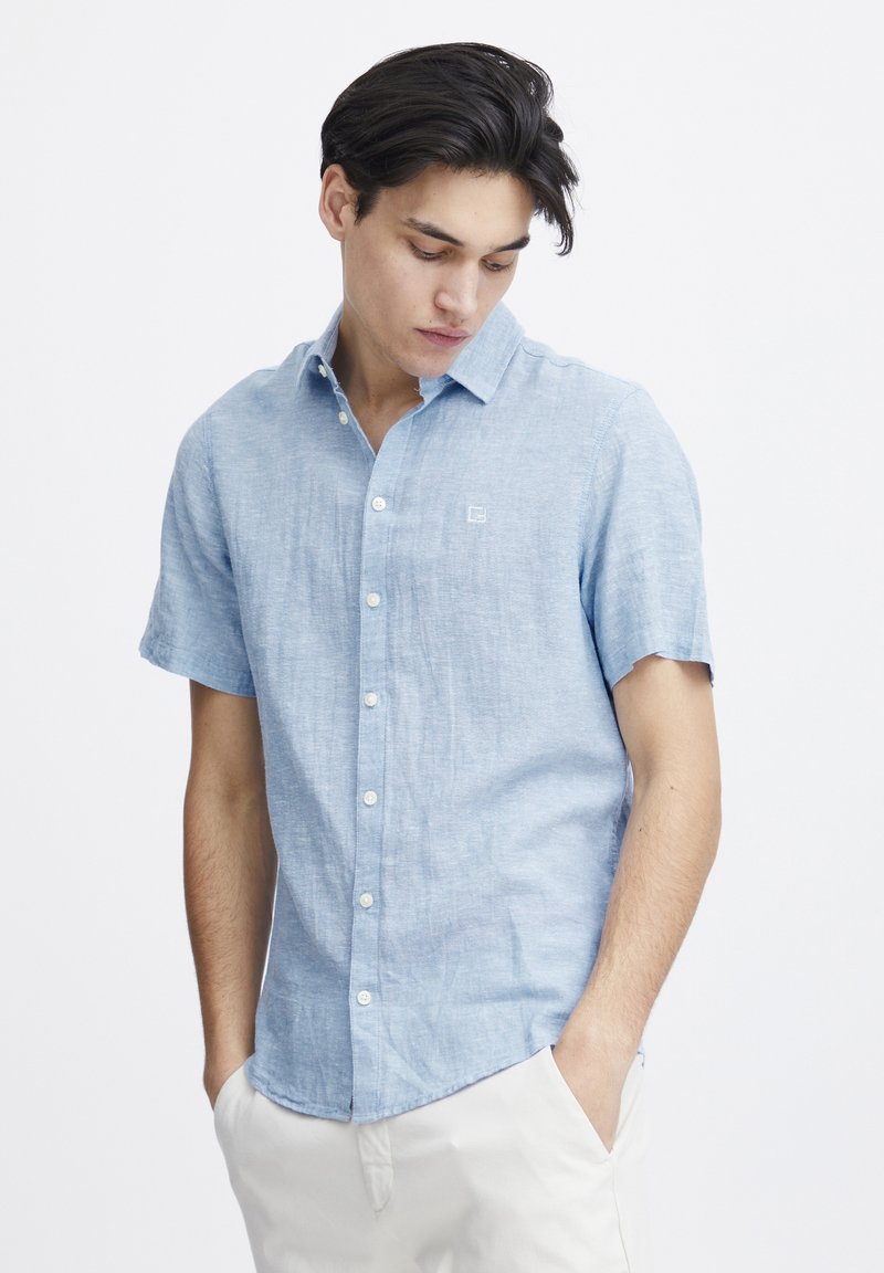 Camicia a maniche corte di colore blu chiaro con colletto a bottoni, realizzata in tessuto traspirante, con un piccolo logo sul lato sinistro del petto.