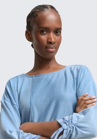Stripet blå topp med bred halslinje og samlede ermer. Stoffet ser mykt og lett ut, med kontrasterende lyse og mørke striper.