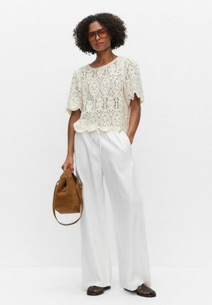 Kvinde med krøllet hår iført mørke solbriller, hvid blonderbluse, brede hvide bukser, sorte vævede sko, bærende på brun ruskindstaske.