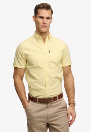 Man draagt een lichtgeel overhemd met korte mouwen en beige broek met een bruine riem, staat met één hand in de zak.