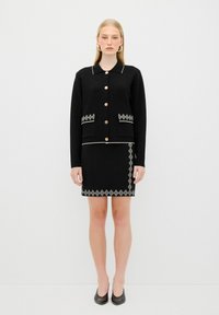 Svart cardigan med guldknappar, vit trim och två framfickor, sammansatt med en svart kjol med en broderad diamantmönster.