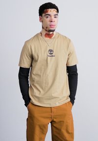 Bežinė trumparankovė marškinėliai su Timberland logotipu, uždėti ant juodų ilgarankovių marškinėlių, derinami su oranžin�ėmis kelnėmis; vientisas fonas.