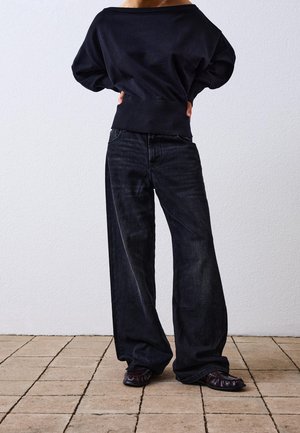 Schwarzer oversized Pullover mit geripptem Saum, kombiniert mit locker sitzenden dunklen Denim-Jeans. Klobige, strukturierte flache Schuhe vervollständigen den Look.