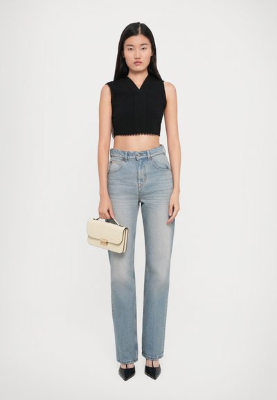 Young woman in sleeveless black crop top, high-waist light blue jeans, black pointed heels, holding cream handbag against plain background.
