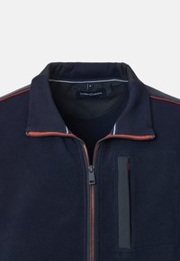 Giacca zip-up blu navy con texture a coste, cerniera rossa e dettagli in piping, colletto alto e taschino verticale sul petto con bordi neri.